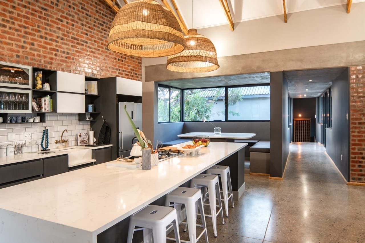 Kitchen with exposed brick walls, a long stone island, banquette seating and rattan pendants