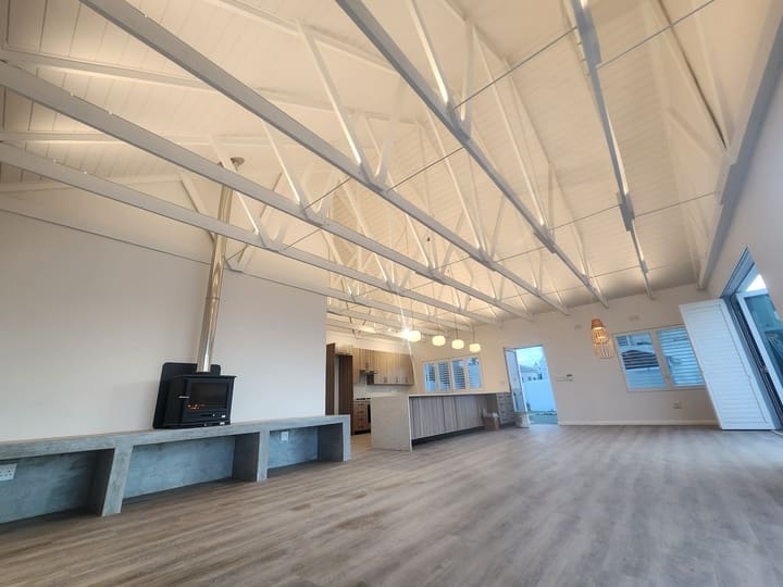 Large open-plan living space with exposed white roof trusses and a wood-burning stove on a concrete plinth